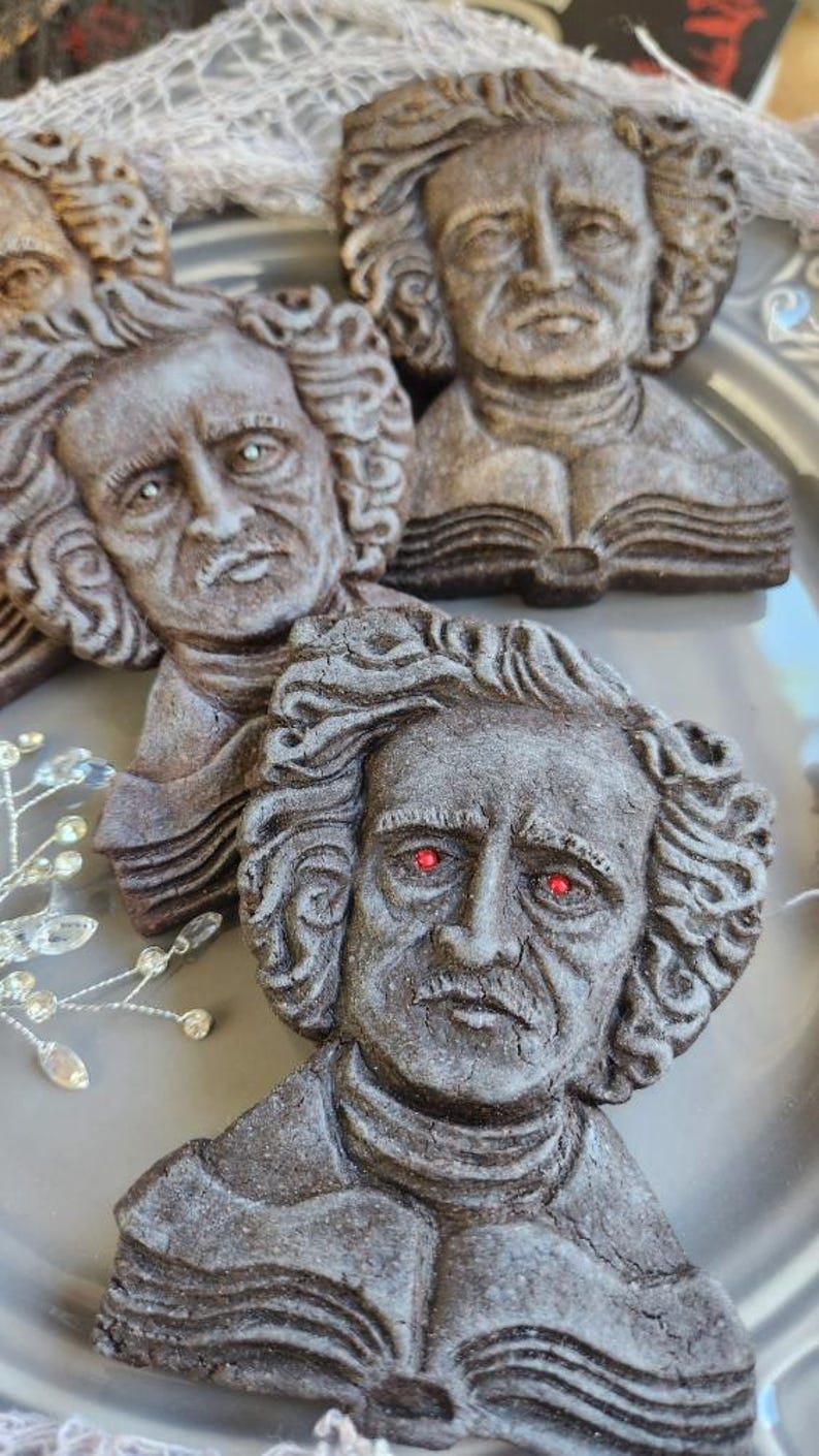 Puede incluir: Primer plano de varias galletas con forma de rostro de Edgar Allan Poe, esculpidas en marr&oacute;n oscuro, con ojos rojos, dispuestas sobre un plato gris. Las galletas est&aacute;n detalladas con pelo rizado y libros abiertos. Se ven algunos detalles decorativos de cristal.