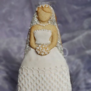 Puede incluir: Una galleta de azúcar blanca decorada para parecer una novia. La galleta tiene un vestido blanco con detalles de encaje, un velo con purpurina plateada y un pequeño ramo de flores blancas.