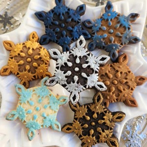 May include: A collection of snowflake-shaped cookies in various colors, including blue, gold, and silver, arranged on a white star-shaped plate. The cookies have intricate details and are decorated with edible paint. A festive holiday treat.