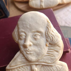 May include: A white sugar cookie shaped like a bust of William Shakespeare. The cookie has a detailed design of Shakespeare's face, hair, and collar.