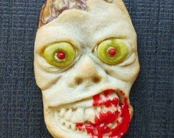 Puede incluir: Una galleta detallada con forma de cara de zombi con glaseado rojo y ojos amarillos. La galleta está hecha para parecer un zombi con una parte de la cabeza que falta.