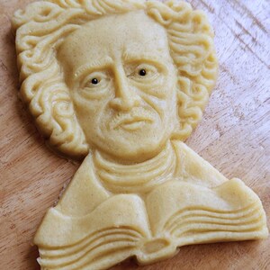 Puede incluir: Una galleta de color claro con forma de busto de hombre con pelo rizado, bigote y un libro. La galleta est&aacute; sobre una superficie de madera. Los rasgos faciales y las p&aacute;ginas del libro son detallados.