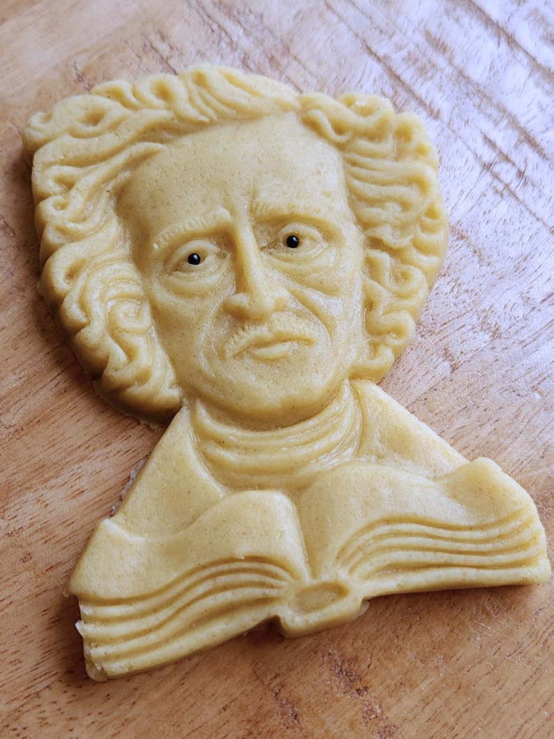 Puede incluir: Una galleta de color claro con forma de busto de hombre con pelo rizado, bigote y un libro. La galleta est&aacute; sobre una superficie de madera. Los rasgos faciales y las p&aacute;ginas del libro son detallados.