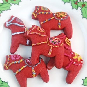 Puede incluir: Cuatro caballos de jengibre rojos decorados con glaseado dorado, azul y blanco en un plato blanco con un borde de acebo verde.