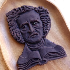 Puede incluir: Una galleta marr&oacute;n oscuro con forma de busto de Edgar Allan Poe, con rasgos faciales detallados y cabello rizado. La galleta descansa sobre una bandeja de madera, destacando el tema literario. Una golosina &uacute;nica.