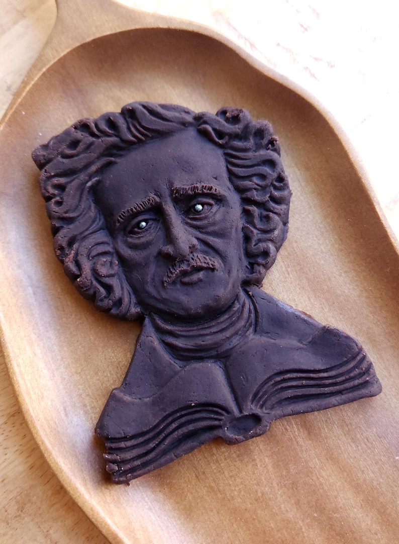 Puede incluir: Una galleta marr&oacute;n oscuro con forma de busto de Edgar Allan Poe, con rasgos faciales detallados y cabello rizado. La galleta descansa sobre una bandeja de madera, destacando el tema literario. Una golosina &uacute;nica.