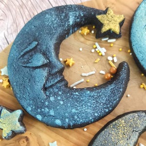 Pode incluir: Dois biscoitos em forma de lua crescente decorados com granulado azul e dourado. Um biscoito tem um rosto e uma estrela dourada no topo. Os biscoitos estão sobre uma superfície de madeira com mais granulado.
