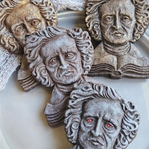 Puede incluir: Una colecci&oacute;n de decoraciones con forma de busto de Edgar Allan Poe. Los bustos son en tonos grises y marrones, con rasgos faciales detallados y cabello rizado. Un busto tiene ojos rojos brillantes. Los bustos est&aacute;n dispuestos sobre un plato gris.