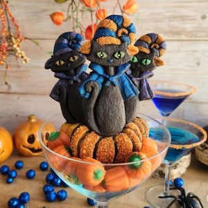 Puede incluir: Una exhibición temática de Halloween con galletas de gato negro con sombreros y capas azules y naranjas, sobre una base de galletas con forma de calabaza. La exhibición está en una copa de martini llena de caramelos de calabaza. Cócteles azules y decoraciones completan la escena festiva.