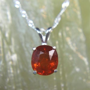 May include: A silver necklace featuring a faceted, oval-shaped, orange-red gemstone pendant. The pendant is held by four prongs and hangs from a delicate, twisted silver chain. The gemstone has a brilliant cut, reflecting light.