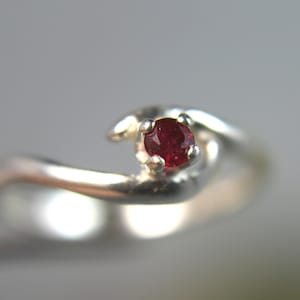 May include: A silver ring featuring a single, round, faceted deep red gemstone set in a four-prong setting. The band has a smooth, polished finish and a curved, bypass-style design that wraps around the central stone, creating a fluid appearance.