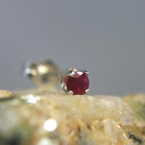 May include: A single stud earring featuring a round, faceted deep red gemstone set in a four-prong silver-toned metal setting. The earring is positioned with the gemstone facing forward, showcasing the vibrant red color and the metallic prongs.