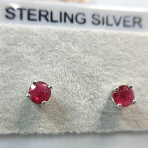 May include: A pair of round, faceted red gemstone stud earrings set in sterling silver prongs. The earrings are mounted on a white display card with the text STERLING SILVER printed in black at the top, indicating the metal composition.