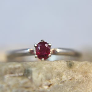 May include: A silver-toned metal ring featuring a single oval-cut deep red gemstone set in a six-prong setting. The band is smooth and polished, showcasing a simple design that highlights the central red stone as the primary focal point.