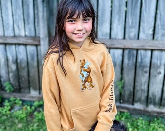 May include: Mustard-colored hoodie with a front pocket and a graphic of a bear hugging a smaller bear. The right sleeve has the text "Yak Island" printed vertically. The hoodie is worn by a person with long brown hair.