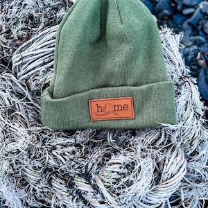 May include: A green knit beanie with a brown leather patch that says "home" with an outline of Alaska on it. The beanie is sitting on a pile of white rope.