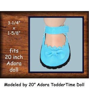 May include: Light blue doll shoes with a bow and scalloped trim. The shoes have a velcro strap and a black sole. The text on the image says the shoes fit a 20-inch Adora doll and are 3-1/4" x 1-5/8".