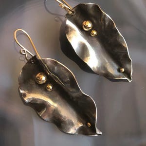 May include: A pair of leaf-shaped earrings. The earrings are made of silver-toned metal with a textured surface and feature small, gold-coloured accents. The ear wires are gold-coloured. The earrings have a nature-inspired design.
