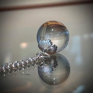 May include: A clear glass orb pendant with a silver chain. The orb has a reflective surface and is attached to a silver pendant with a shell-like design. The chain is made of small, interconnected silver beads. The pendant is displayed on a reflective surface.