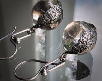 Può includere: Un paio di orecchini pendenti con perle di vetro sferiche trasparenti parzialmente racchiuse in cappucci di metallo color argento ossidato scuro. I cappucci in metallo sono decorati con un motivo floreale dettagliato di margherite e foglie.