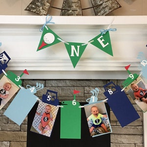 May include: A golf-themed birthday banner with blue, green, and red paper cutouts. The banner has a golf cart design with a number on each cutout. The banner also has a golf ball and the word 