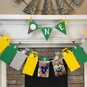 May include: A green, yellow, and gray banner with photos of a baby boy at different ages. The banner has the numbers 1 through 12 and the letters N, B, and E. The banner is decorated with golf-themed images.