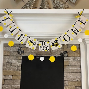 May include: A white and yellow banner with black bee illustrations and the text "Fun to Bee One". The banner is strung across a white fireplace mantel.