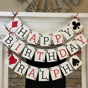 May include: A white and red banner with black letters that reads "Happy Birthday Ralph" with playing card symbols on each card. The banner is strung up on a fireplace mantel.