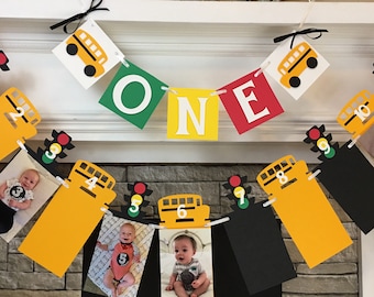 May include: A yellow and black school bus themed first birthday banner with photos of a baby boy. The banner includes the word "ONE" in green, yellow, and red letters. Each photo is attached to a yellow school bus with a number from 1 to 10. Each bus also has a traffic light with red, yellow, and green lights.