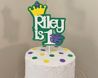 Puede incluir: Un pastel blanco con lunares verdes, amarillos y morados. Un adorno para pastel con fondo verde, letras blancas que dicen "Riley is 1", una corona amarilla y una máscara morada.
