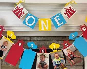 May include: A colourful banner with red, yellow, blue and white paper cutouts. The banner says "ONE" and has numbers 1 through 10 with pictures of a baby in each cutout. The banner is hanging from a white fireplace mantel.