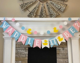 May include: A pink and blue gender reveal banner with the text "Waddle It Be?" and yellow rubber duck decorations. The banner is strung across a white mantelpiece with white and blue polka dots.