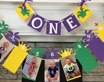 May include: A Mardi Gras themed first birthday banner with purple, green, and gold colors. The banner has a large "ONE" in the center and 12 smaller flags with numbers 1-12 and photos of a baby.
