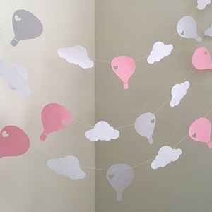 Puede incluir: Una guirnalda de globos de papel rosa y gris y nubes de papel blanco. Los globos tienen un peque&ntilde;o coraz&oacute;n recortado en el centro.