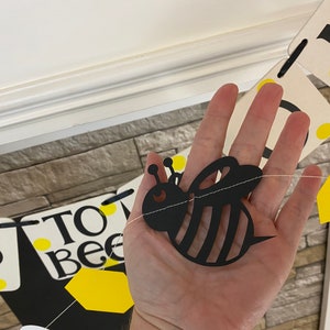 May include: A black and yellow bee cutout with a white string attached to it. The bee is hanging from a banner that says "To Bee".