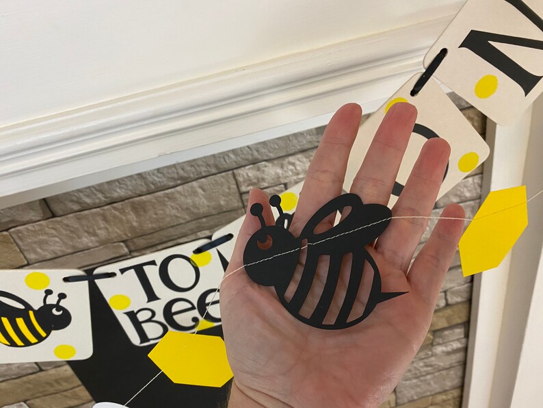 May include: A black and yellow bee cutout with a white string attached to it. The bee is hanging from a banner that says "To Bee".
