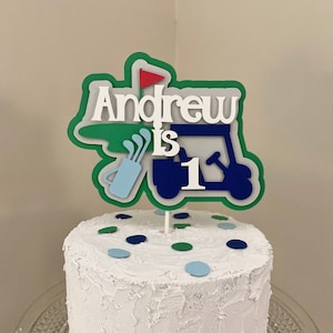 May include: A white cake with blue and green sprinkles topped with a green, blue, and red golf-themed cake topper that says "Andrew is 1".