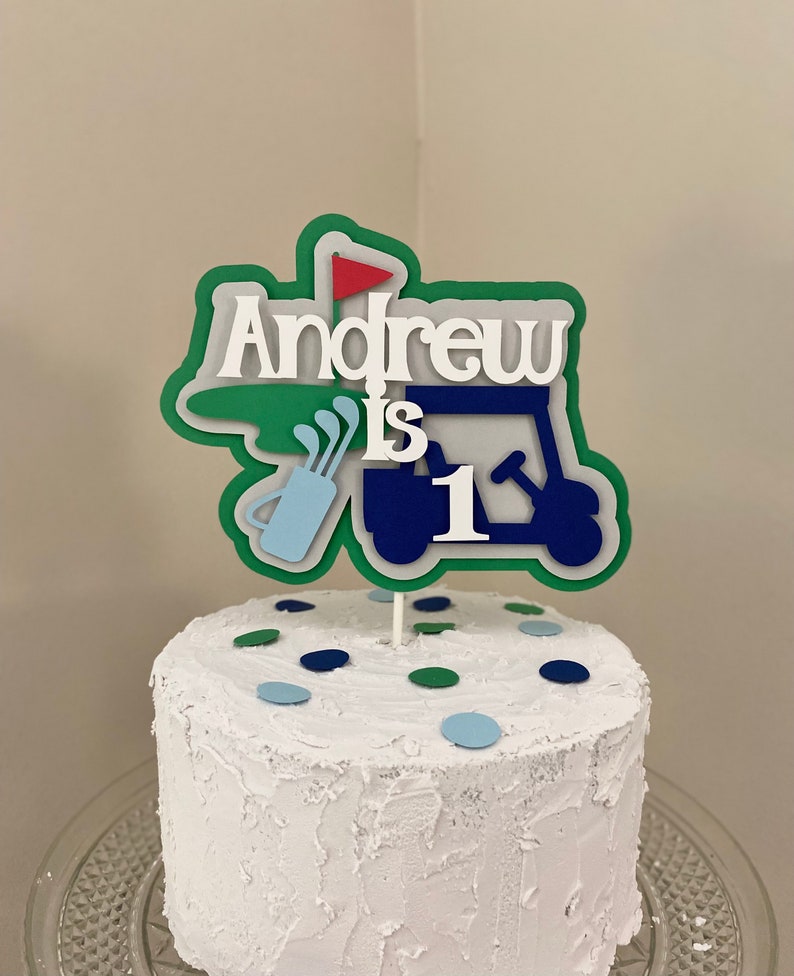 May include: A white cake with blue and green sprinkles topped with a green, blue, and red golf-themed cake topper that says "Andrew is 1".