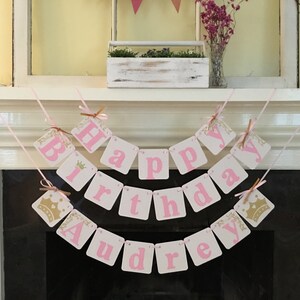 May include: Pink and gold banner with the words "Happy Birthday Audrey". The banner is made of cardstock and has a crown design on each end.