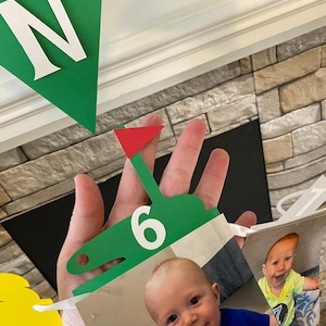 May include: A green and red golf-themed banner with a white number six, a white letter N, and a photo of a baby boy. The banner is made of paper and is decorated with golf clubs and a golf ball.