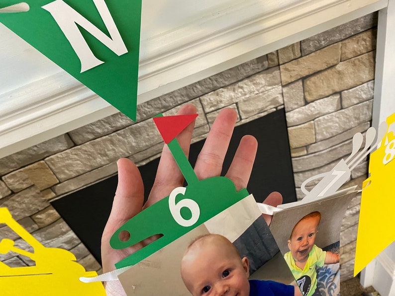 May include: A green and red golf-themed banner with a white number six, a white letter N, and a photo of a baby boy. The banner is made of paper and is decorated with golf clubs and a golf ball.