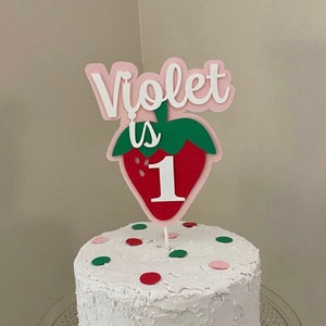 May include: A white cake with red and green sprinkles topped with a pink and green strawberry cake topper that says "Violet is 1".