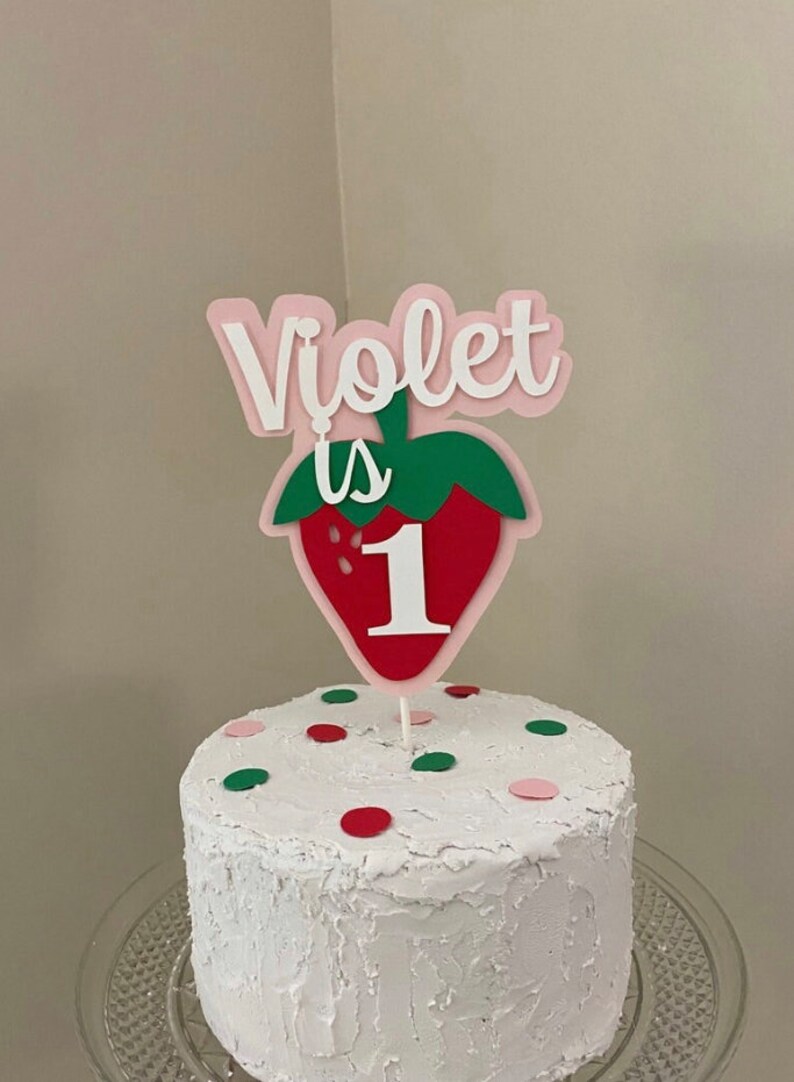 May include: A white cake with red and green sprinkles topped with a pink and green strawberry cake topper that says "Violet is 1".