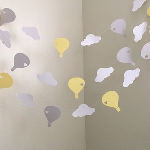 Puede incluir: Una guirlande de globos de papel amarillo y gris en forma de globos aerost&aacute;ticos y nubes. Cada globo tiene un peque&ntilde;o coraz&oacute;n recortado. La guirlande est&aacute; enhebrada con hilo blanco.