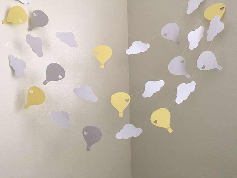 Puede incluir: Una guirlande de globos de papel amarillo y gris en forma de globos aerost&aacute;ticos y nubes. Cada globo tiene un peque&ntilde;o coraz&oacute;n recortado. La guirlande est&aacute; enhebrada con hilo blanco.
