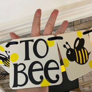 May include: A white banner with black text that reads "To Bee" with yellow polka dots and black bees. The banner is strung together with black ribbon.