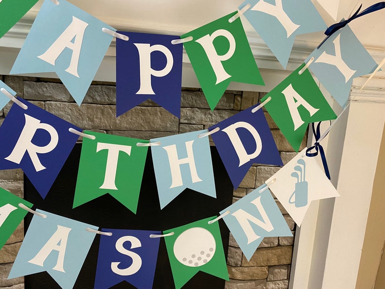 May include: Happy Birthday banner with blue, green, and white flags. The flags have the letters "Happy Birthday" and a golf ball and golf clubs.