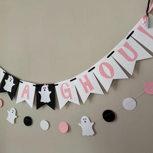 以下が含まれることがあります： ピンクと黒の文字と幽霊の切り抜きが施された「It's a ghoul」の横断幕。横断幕は白い紐でつなげられており、ピンクと黒の円形が飾られています。