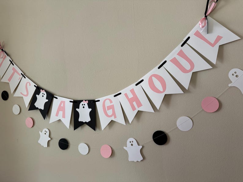 以下が含まれることがあります： ピンクと黒の文字と幽霊の切り抜きが施された「It's a ghoul」の横断幕。横断幕は白い紐でつなげられており、ピンクと黒の円形が飾られています。