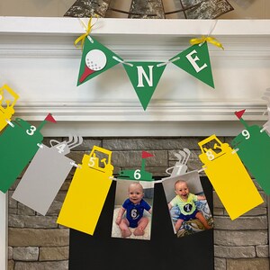 May include: A green and yellow construction vehicle themed banner with photos of a baby boy. The banner has the word "ONE" in white letters on a green triangle. The banner is strung with clips and has numbers 1 through 10 on each section.